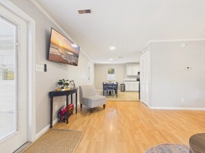 A spacious living area with hardwood flooring, a comfortable armchair, a dining table, and a kitchen in the background. - `The Kelly` - The Cottage Collection (Virginia Beach)