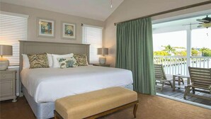 Une chambre d’hôtel avec un grand lit, deux lampes de chevet, un banc, un ventilateur de plafond et une vue sur un balcon aménagé avec du mobilier d’extérieur.