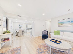 A modern living room with a white sofa, a coffee table, and a dining area in the background. - `The Greyson` - The Cottage Collection (Virginia Beach)