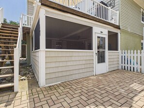 A covered patio area with a wooden deck, a white door, and a white picket fence. - `The Greyson` - The Cottage Collection (Virginia Beach)