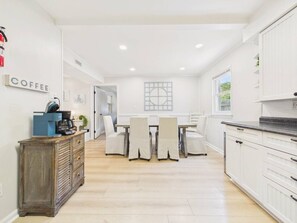 A modern kitchen with white cabinetry, a coffee station, and a dining area with chairs and a table. - `The Greyson` - The Cottage Collection (Virginia Beach)