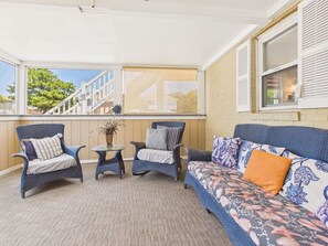 A covered outdoor seating area with wicker furniture, a small table, and a sofa with patterned cushions. - `The Greyson` - The Cottage Collection (Virginia Beach)