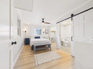 A bedroom with a bed, a ceiling fan, a bedside table, a bench, and a view into the bathroom. - `The Greyson` - The Cottage Collection (Virginia Beach)