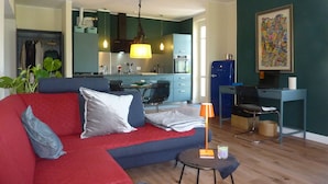 En stue med sofa, lille bord, lampe og et køkken i baggrunden.