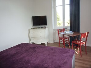 Una habitación con cama, escritorio y silla, televisor en una cómoda, y ventana con cortinas.