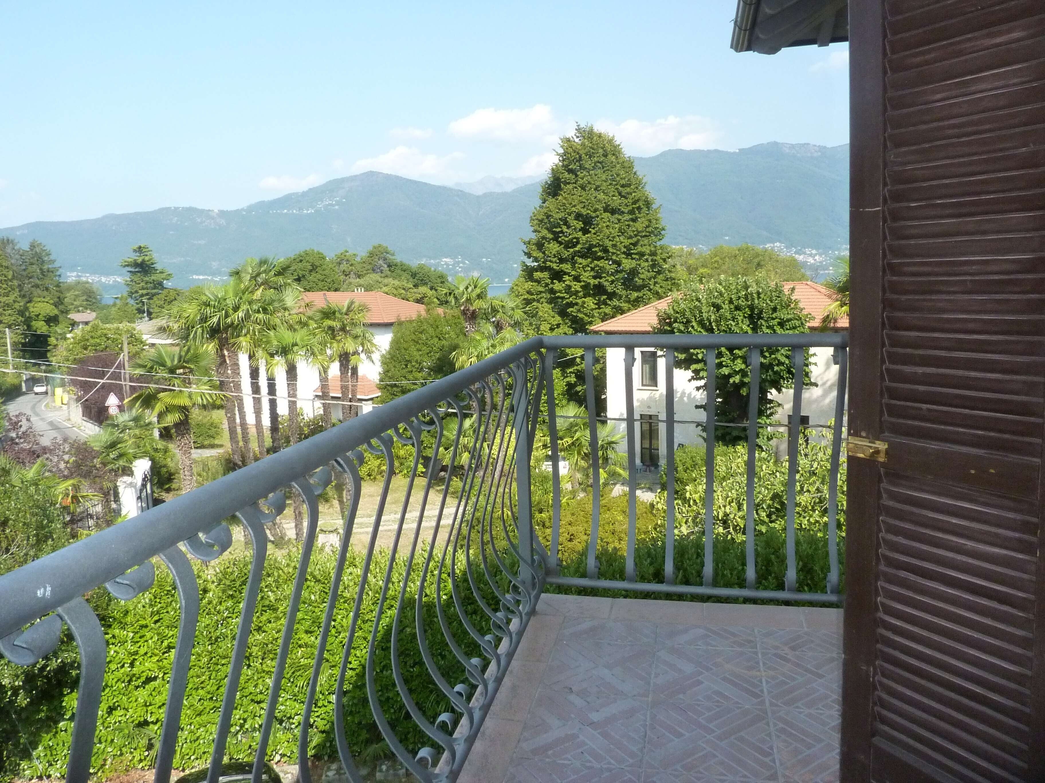 Un balcon doté d’une balustrade en métal, donnant sur un quartier résidentiel avec des maisons, de la verdure et, au loin, des montagnes.
