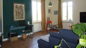 En stue med en blå sofa, et skrivebord, en stol, en stor grøn plante og et maleri på væggen.