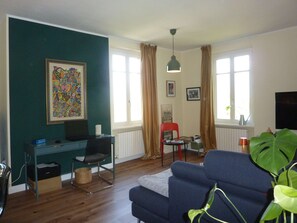 Ein Wohnzimmer mit einem blauen Sofa, einem Schreibtisch, einem Sessel, einer großen Zimmerpflanze und einem Bild an der Wand.
