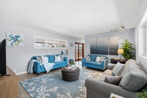 A living room with two sofas, a coffee table, and a rug. There are paintings on the walls and a plant on the right. - Family‑Friendly Tropical Beach Home w/ Private Pool ☀️ (Indian Harbour Beach)