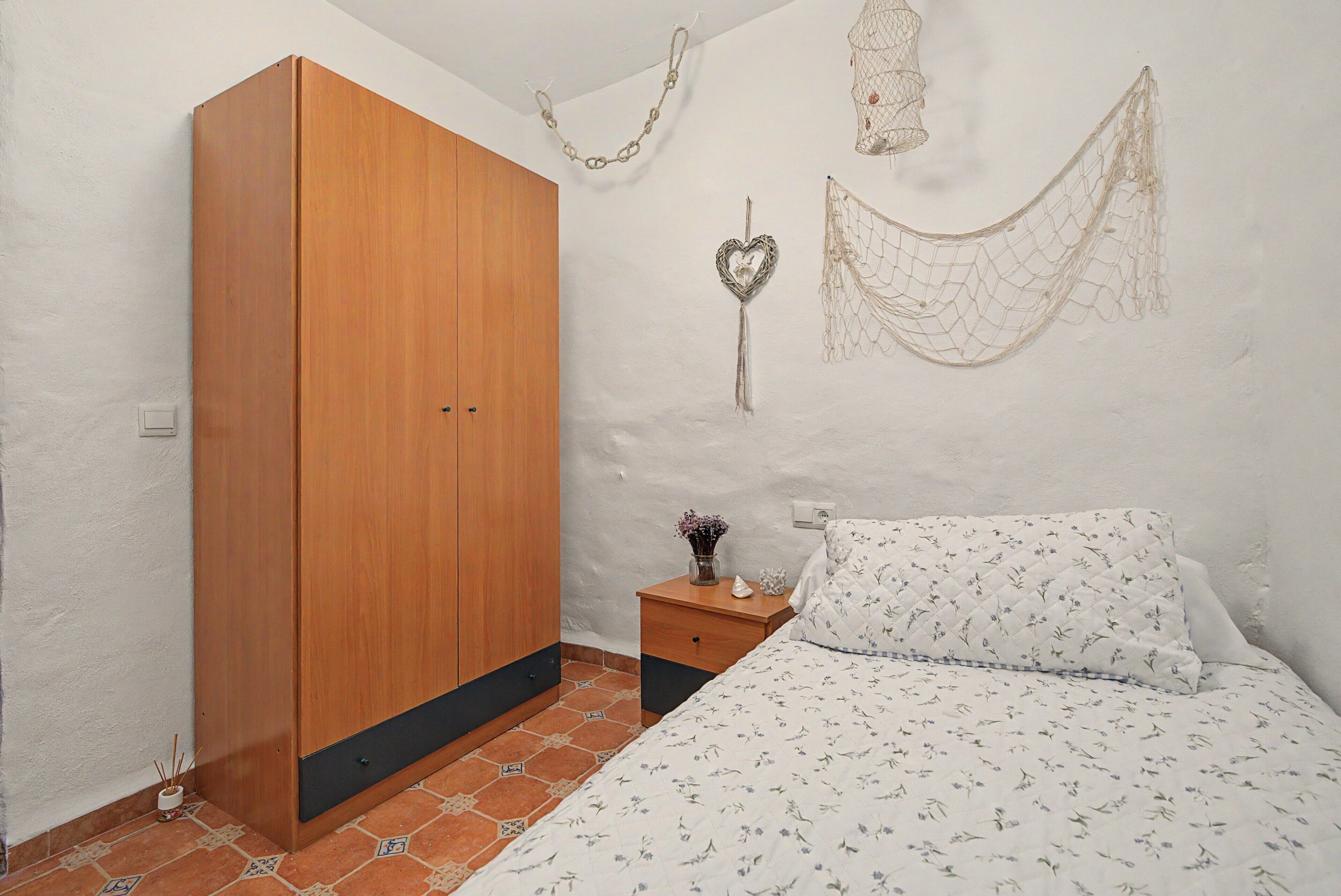 Un dormitorio con un armario de madera, una mesita de noche con un florero y un objeto decorativo, y una cama con edredón estampado de flores.