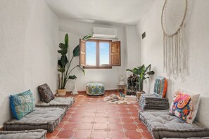 A cozy room with a tiled floor, a window with wooden shutters, and various cushions and plants.