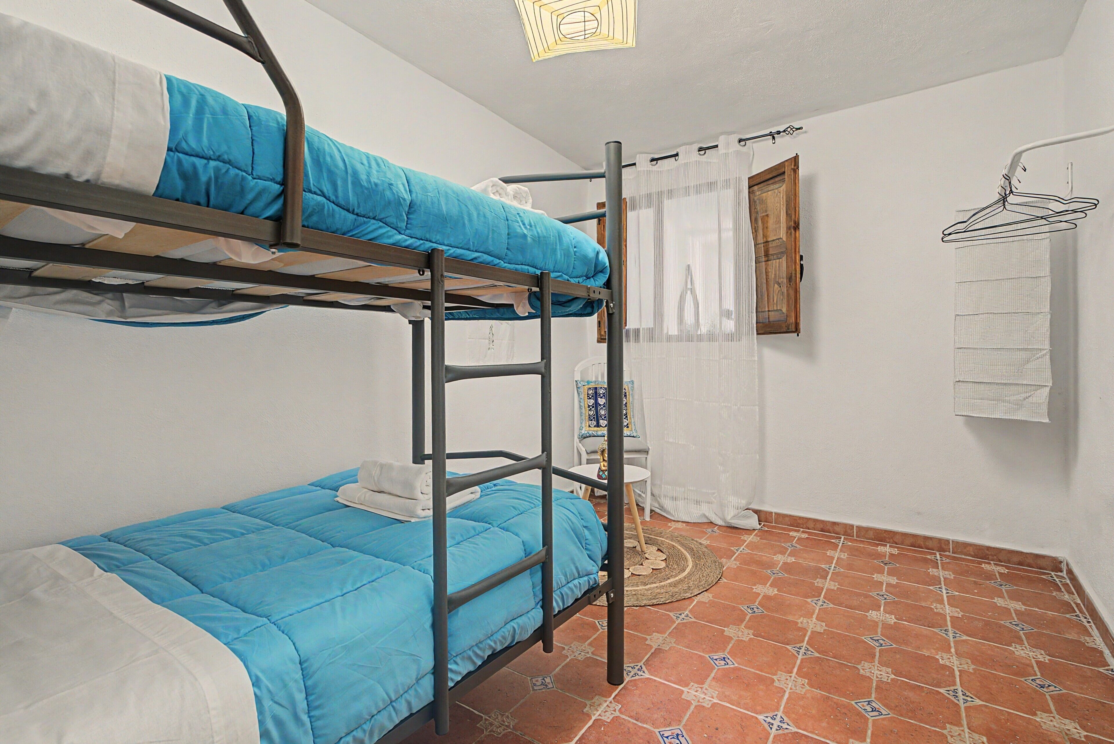 Quarto de dormitório com beliches, uma mesa pequena, uma cadeira, janela com cortinas e um porta-toalhas.
