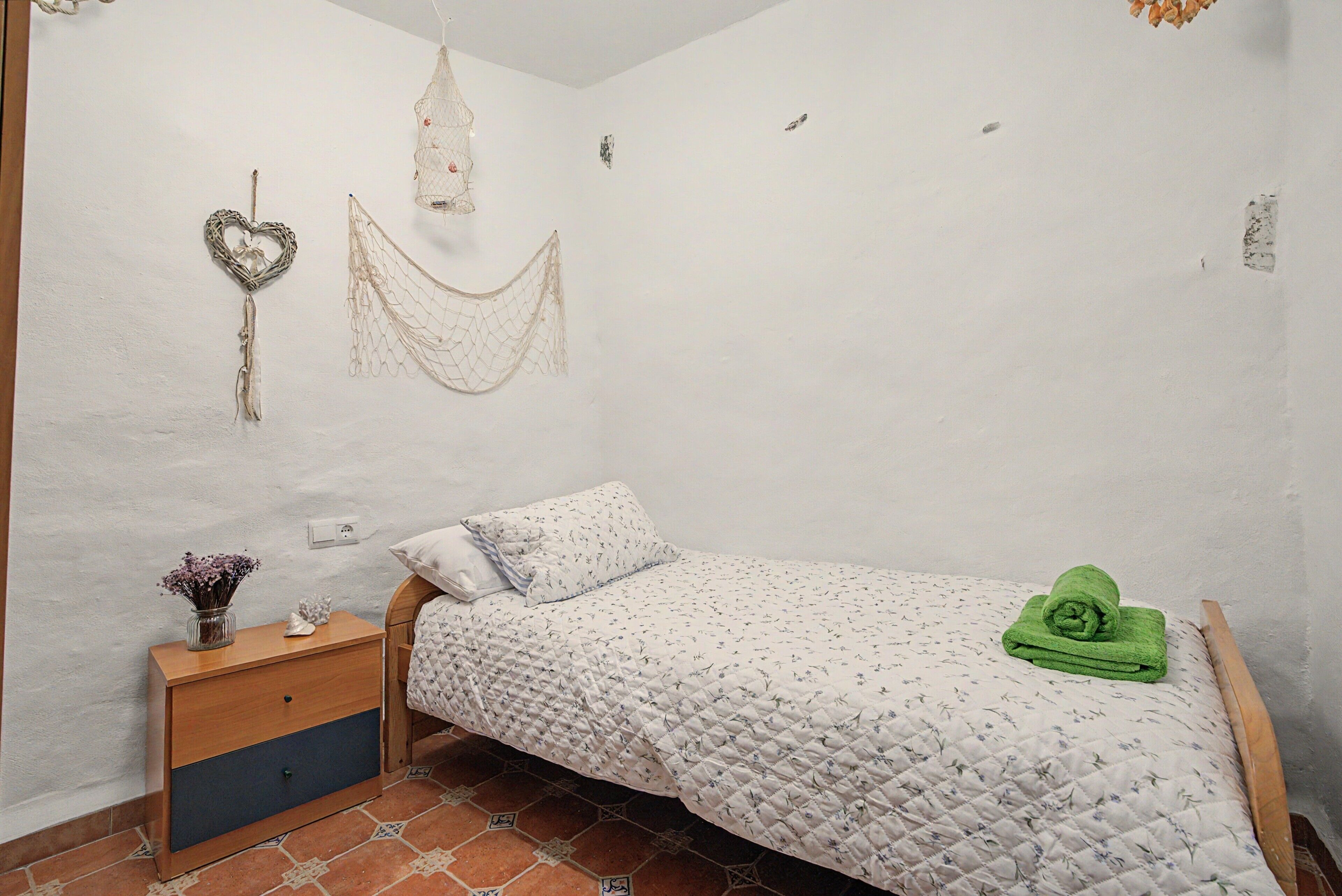 Un dormitorio pequeño y acogedor con una cama individual, una mesita de noche, una hamaca decorativa y un jarrón pequeño con flores.