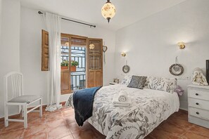 A bedroom with a bed, a chair, a nightstand, a wall-mounted lamp, and a window with curtains.