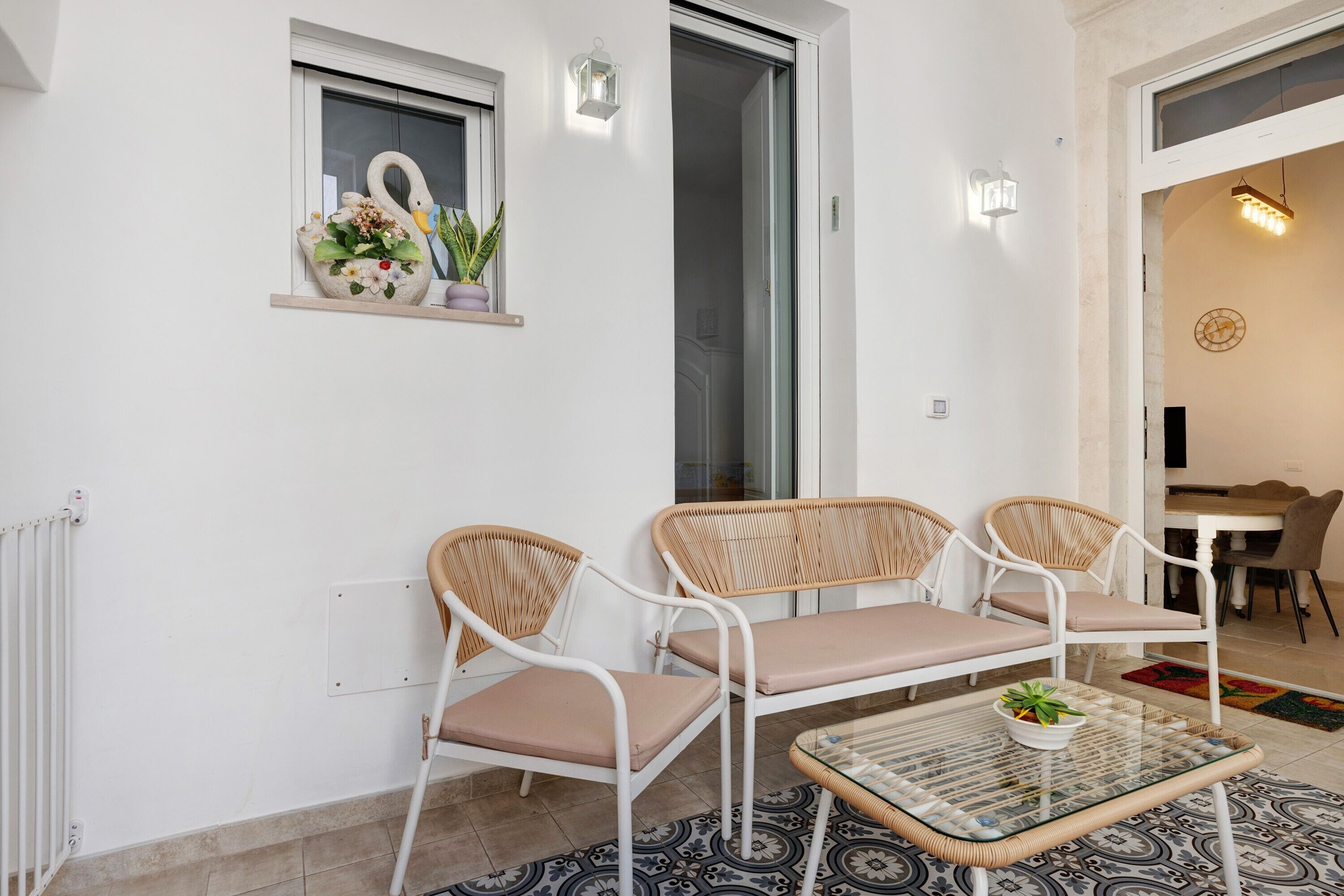 Une pièce avec un mur blanc, une fenêtre ornée d’un cygne décoratif, une petite étagère avec des fleurs, une table en verre et une chaise en osier, ainsi qu’une porte donnant sur une autre pièce.