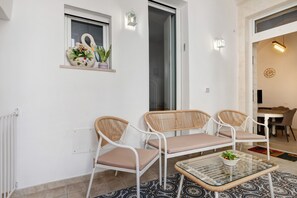 Une pièce avec un mur blanc, une fenêtre ornée d’un cygne décoratif, une petite étagère avec des fleurs, une table en verre et une chaise en osier, ainsi qu’une porte donnant sur une autre pièce.