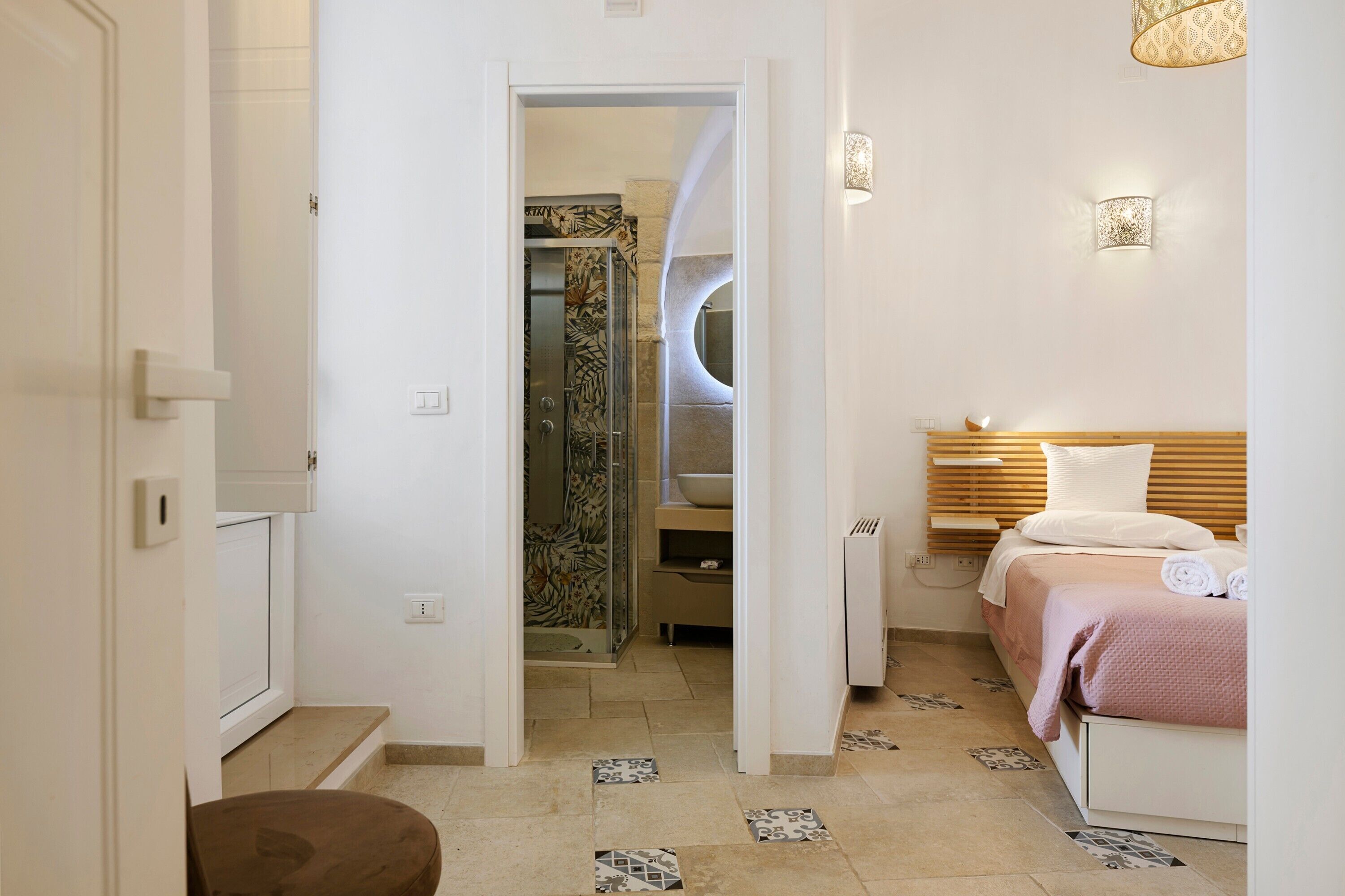Une chambre d’hôtel avec une douche, un lit doté d’une tête de lit en bois, une table de chevet et un petit tabouret.