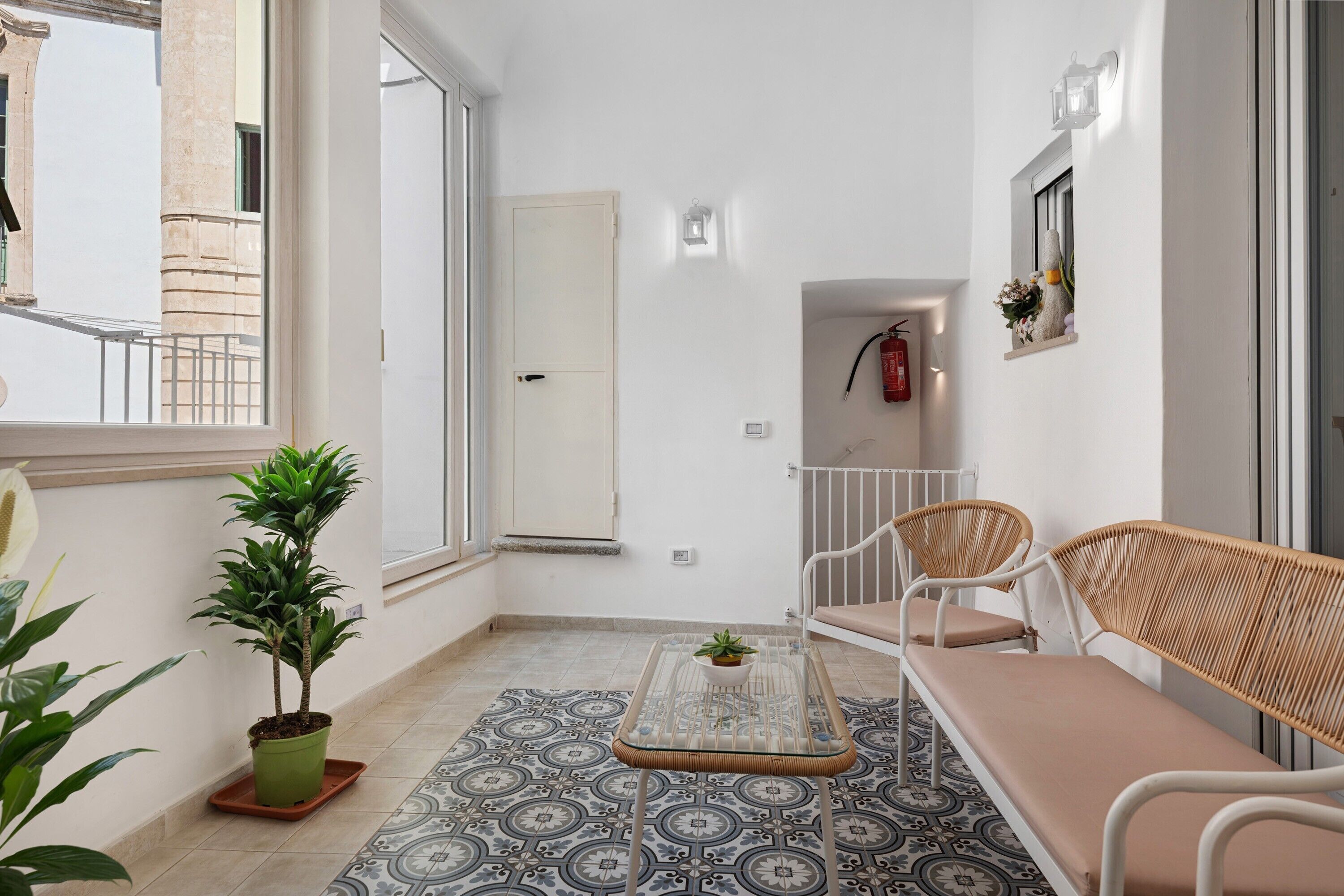 Un balcon avec une balustrade blanche, un pot de fleurs, une table à plateau en verre, des meubles en osier et des carreaux de sol à motifs.