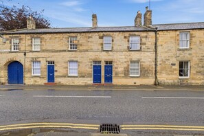 A two-story stone building with multiple doors and windows, a paved road in front, and a drainage grate. - The Old Forge - North East Escapes (Warkworth)