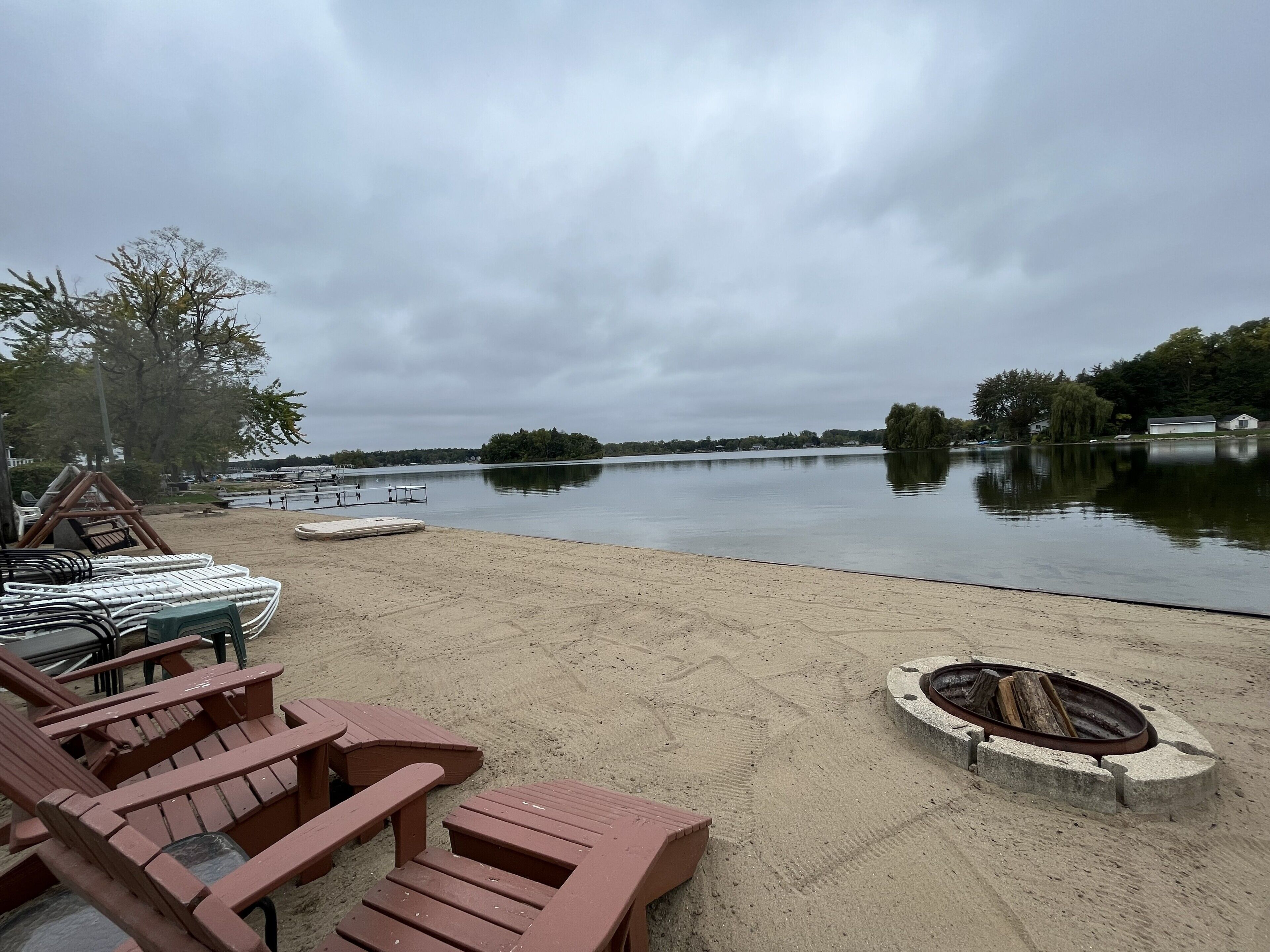 Een gebied aan een meer met een zandstrand, houten ligstoelen en een vuurkuil.