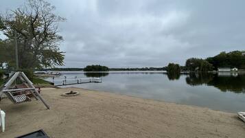 A lakeside view with a sandy beach, a swing set, and a dock extending into the water.