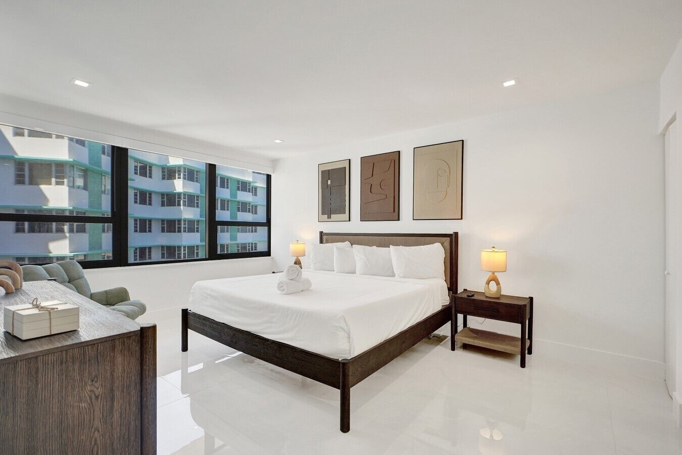 A modern hotel room with a large bed, bedside lamps, a wooden nightstand, and a view of a building through large windows.