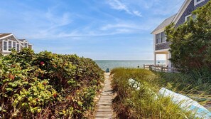 A coastal pathway leading to a beach with a wooden fence and dense foliage on one side and a building on the other.