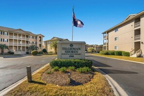 A sign for 'TILGHMAN SHORES Ocean & Golf Resort' with a flag, indicating a multi-story building with balconies and a landscaped area in front. - Tilghman Shores I6- 2 Bedroom- Pool, Hot Tub (North Myrtle Beach)