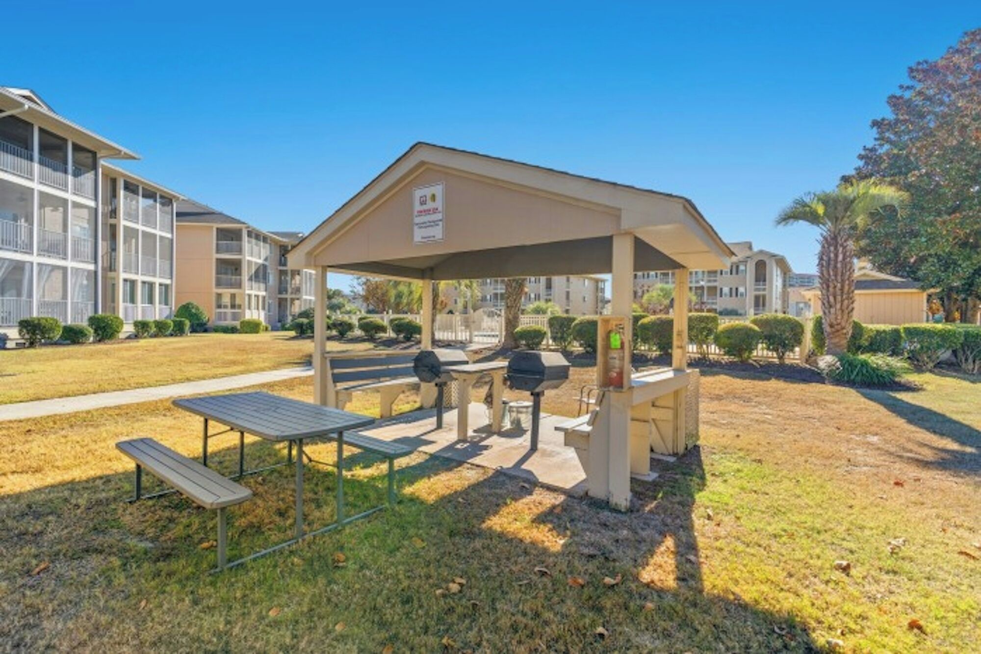Un espacio al aire libre techado con mesas de picnic y parrillas, rodeado de un césped cuidado y edificios residenciales al fondo.