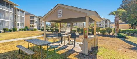 Un espacio al aire libre techado con mesas de picnic y parrillas, rodeado de un césped cuidado y edificios residenciales al fondo.
