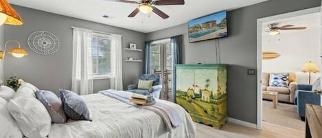 Un dormitorio con cama, ventilador de techo, ventana, un cuadro y puerta que da a otra habitación.