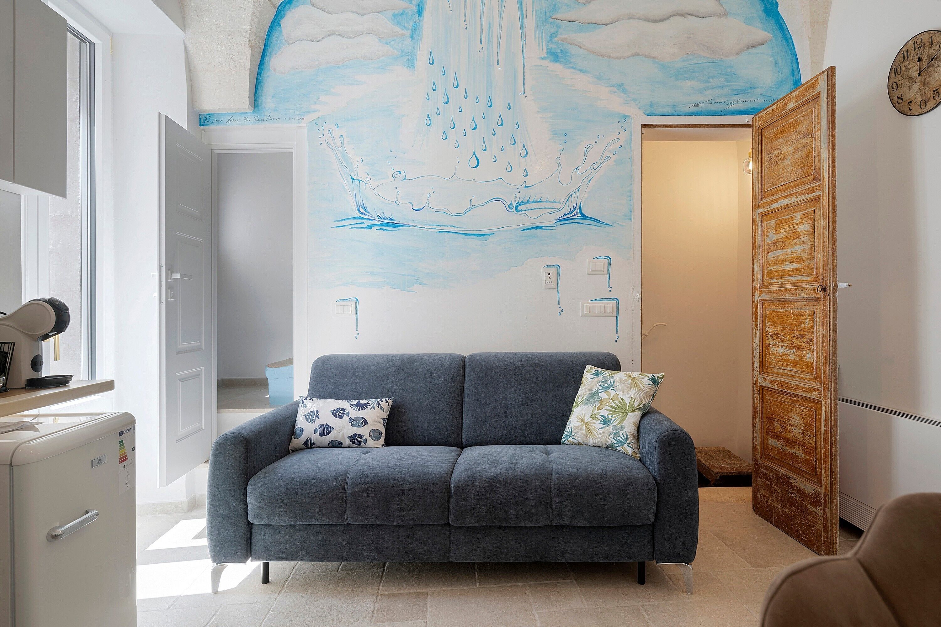 Una habitación con un sofá gris, un mural en la pared, una puerta de madera y un armario blanco.