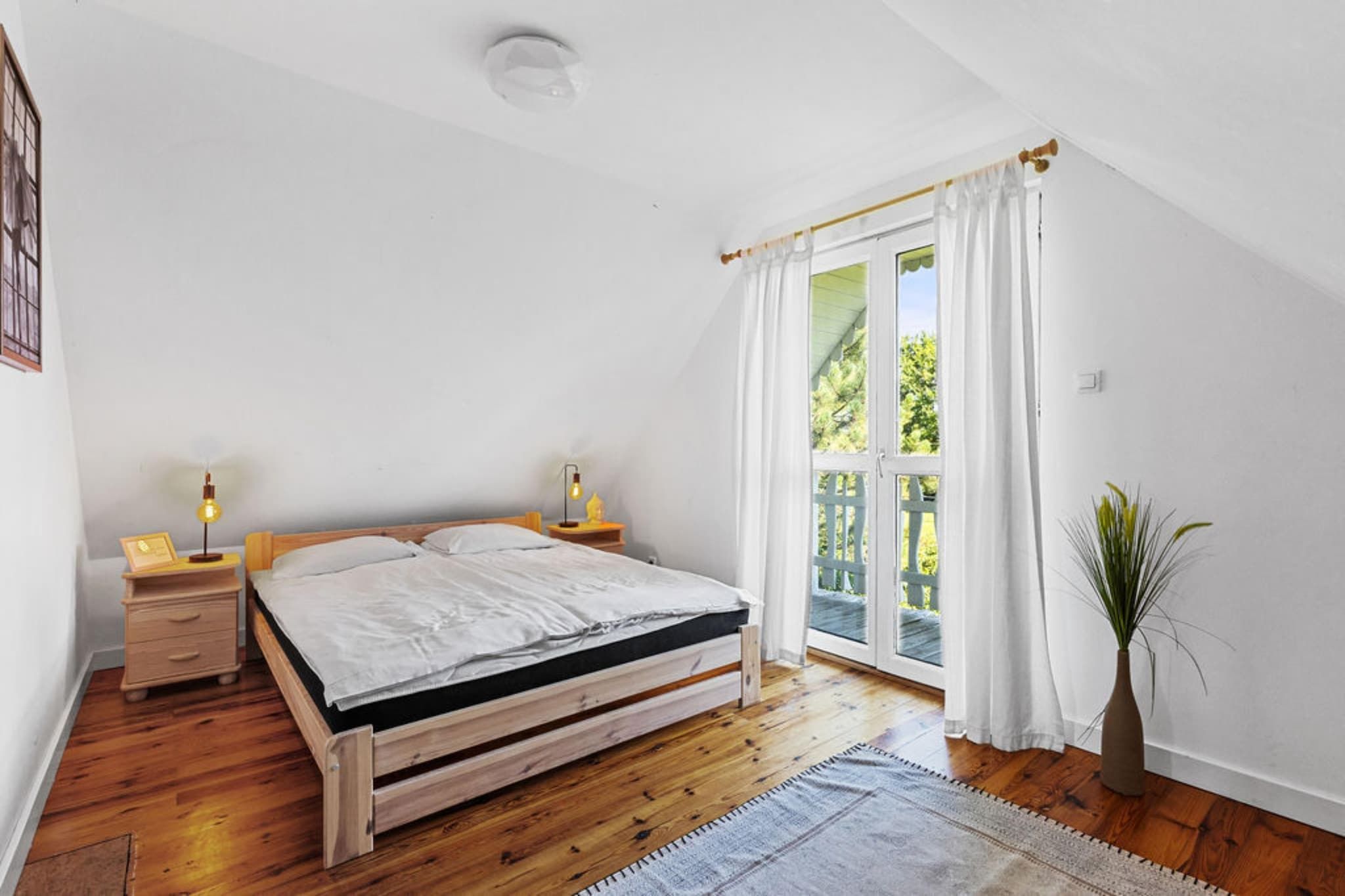 A bedroom with a wooden bed, two bedside tables with lamps, a large window with curtains, and a plant.