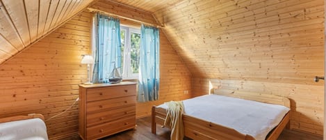 Una habitación de madera con techo inclinado, una cama, un aparador y una ventana con cortinas.