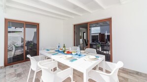 Un comedor con una mesa blanca preparada para cuatro personas, rodeada de sillas blancas, con vista al patio exterior.