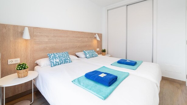 Habitación de hotel con cabecera de madera, dos camas, una mesita de noche con una planta y una lámpara de pared.