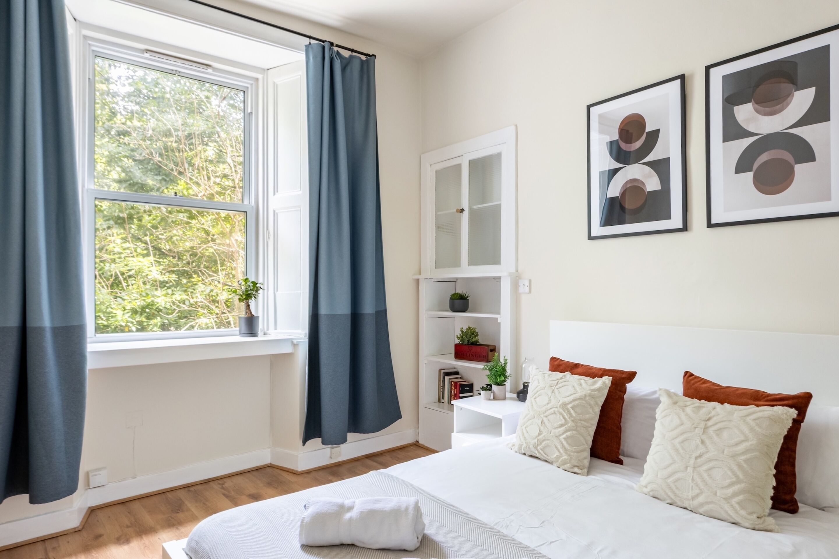 Une chambre à coucher comprenant un lit, une table de chevet, une fenêtre avec des rideaux, une bibliothèque et deux tableaux encadrés accrochés au mur.