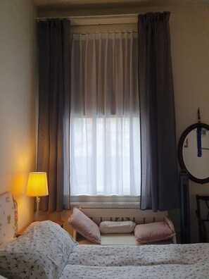 Ein Schlafzimmer mit einem Bett, einer Nachttischlampe, einem Fenster mit Vorhängen, einem Spiegel und einer Heizung.