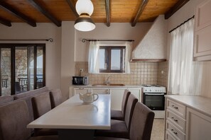 A kitchen with a dining table, chairs, a microwave, a stove, and a window with curtains. - Villa Koule - Three Bedroom Villa, Sleeps 6 (Kalami)