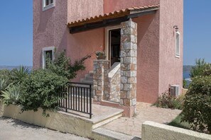 A pink building with a stone staircase leading to a door, surrounded by greenery and a clear view of the sea. - Villa Koule - Three Bedroom Villa, Sleeps 6 (Kalami)