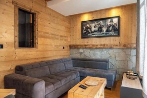 A wooden-paneled room with a gray sectional sofa, a stone wall, and a framed picture above the sofa. - Le Nid des Montagnes - Wood, Stone, and Serenity (Gérardmer)