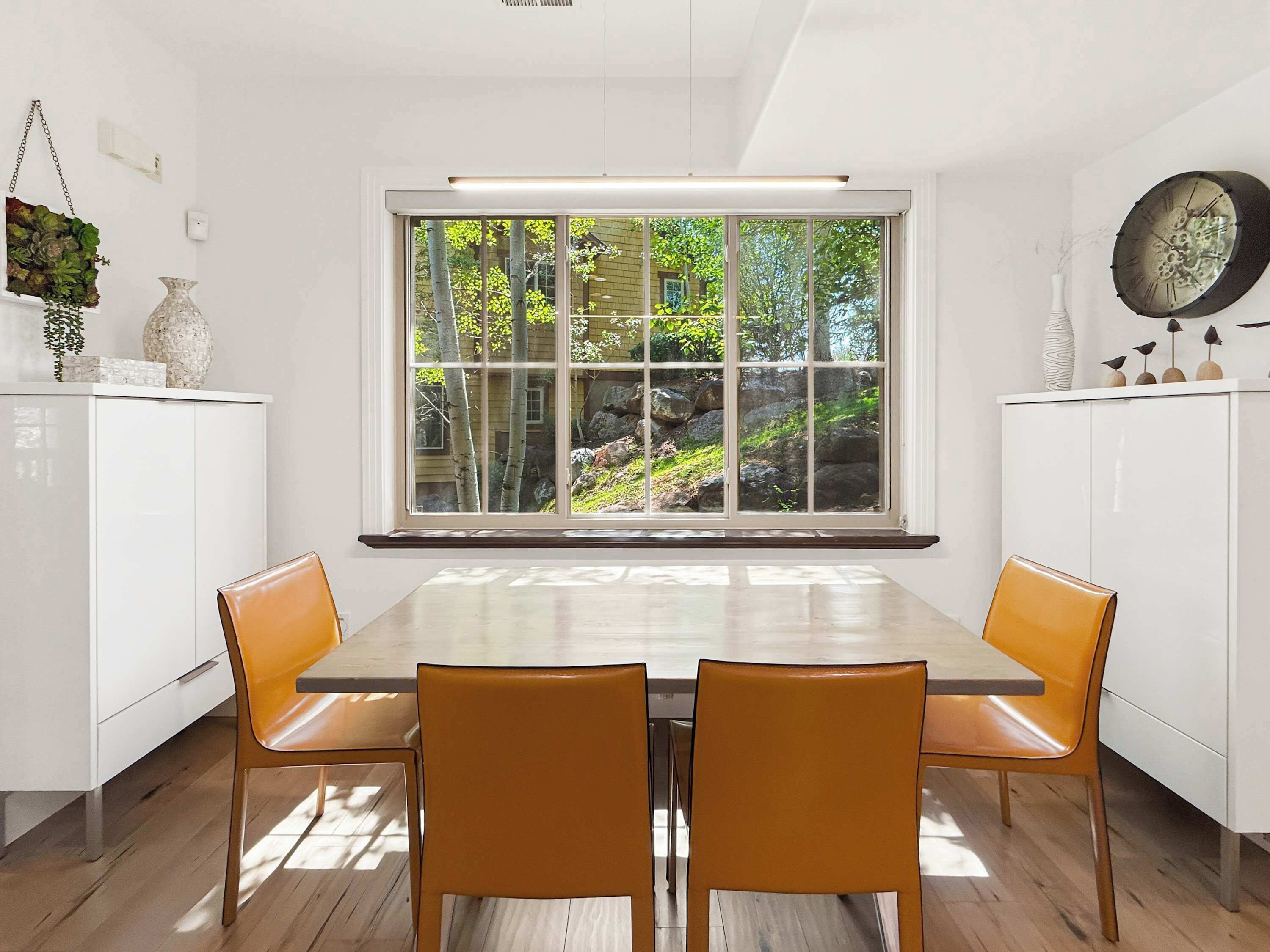 A dining area with a large window, white cabinetry, and two orange chairs.