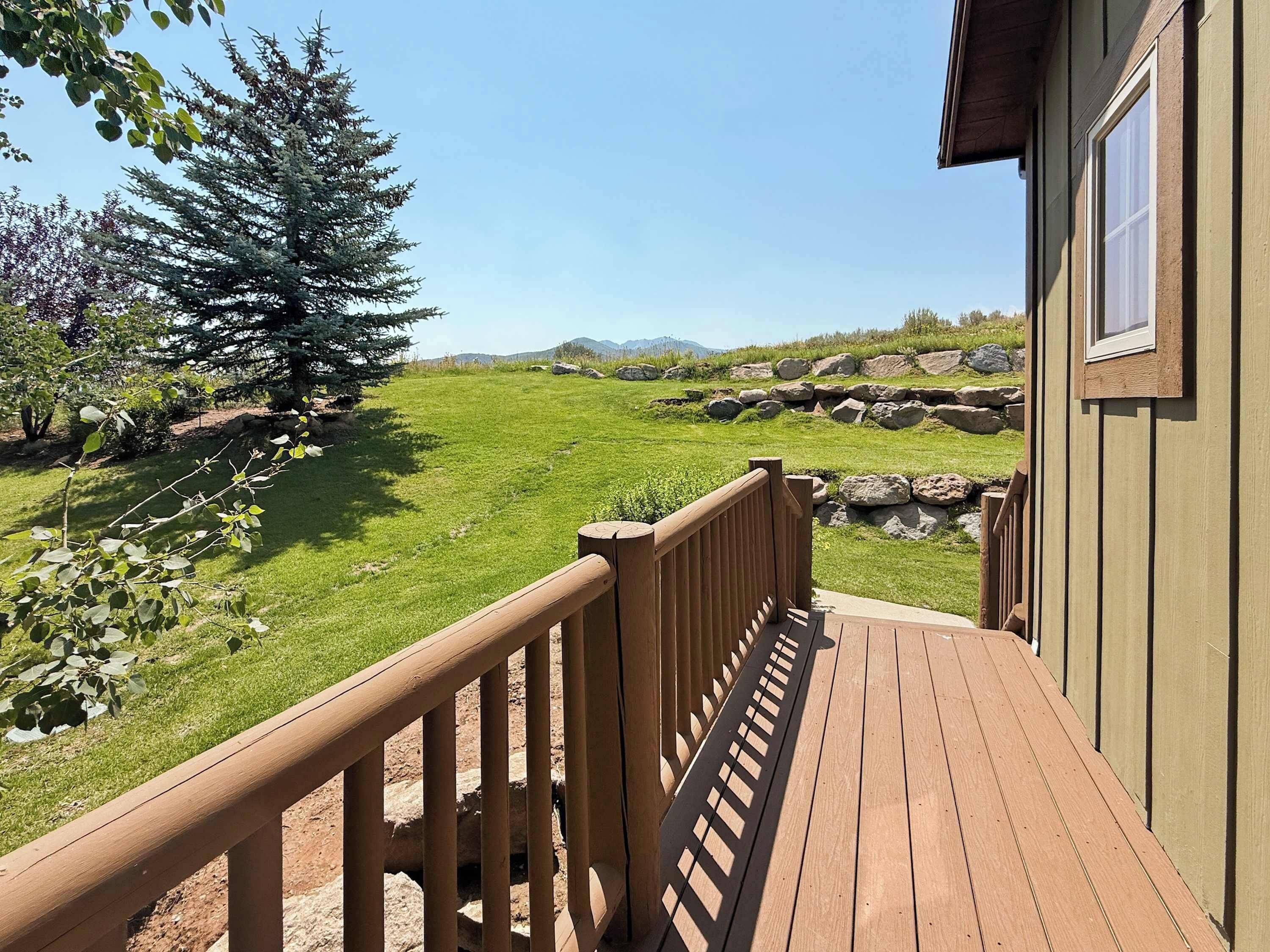 A wooden deck leading to a building with a view of a grassy field and a large tree.