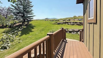 A wooden deck leading to a building with a view of a grassy field and a large tree.