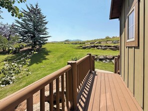 A wooden deck leading to a building with a view of a grassy field and a large tree.