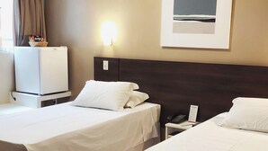 A hotel room with two beds, a small desk, a refrigerator, a wall-mounted light, and a framed picture.