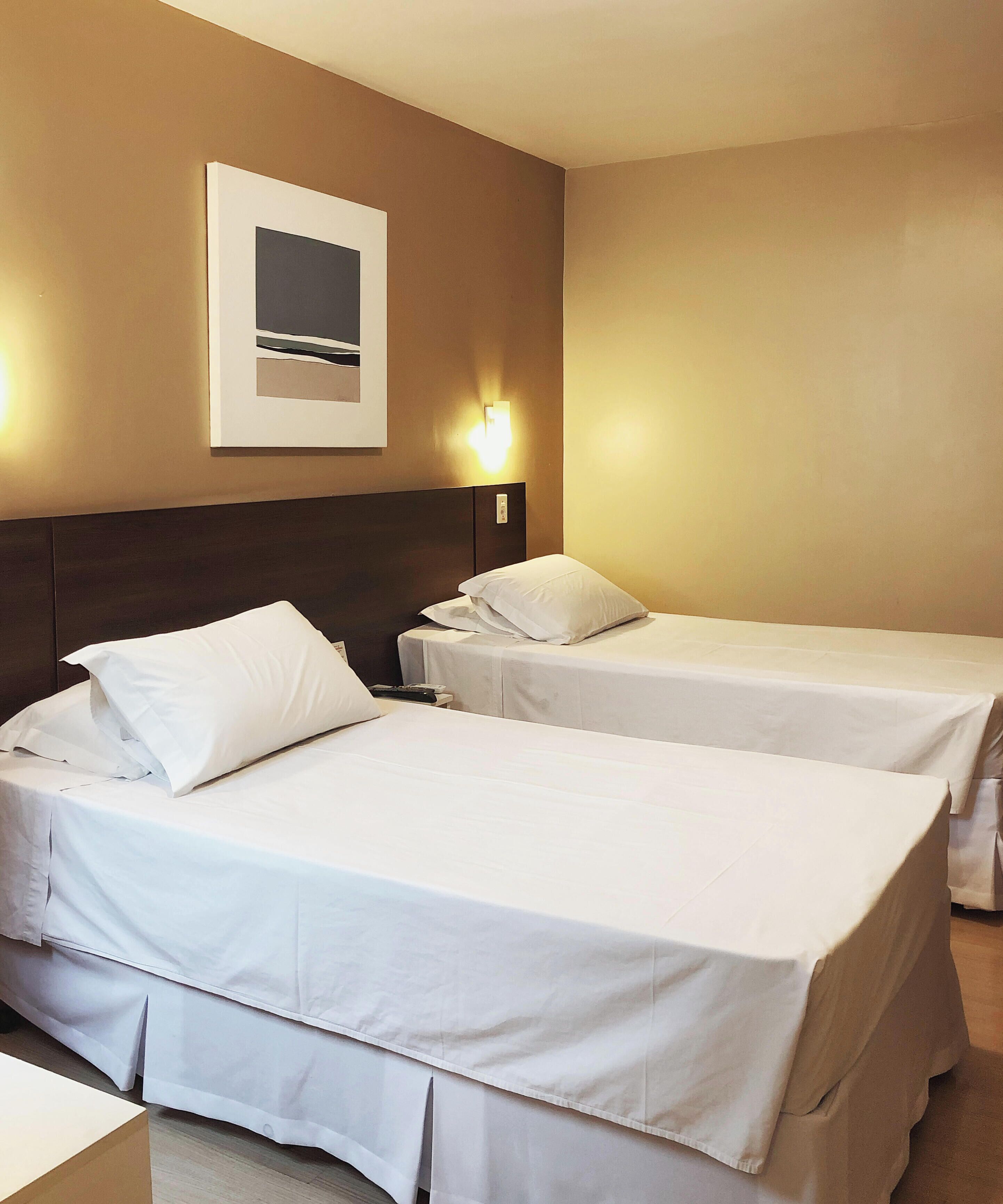 Ett hotellrum med två separata sängar, en sänggavel, en väggmonterad lampa och en inramad tavla.