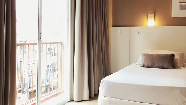 A hotel room with a bed, a nightstand, a wall-mounted light, and a view of the city through a large window.