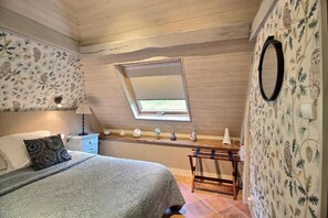 Ein Schlafzimmer mit schrägen Wänden, einem Oberlicht, einem Bett mit gemusterten Betttextilien, einem Nachttisch mit Lampe und einem Regal mit Dekoration.