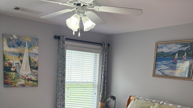 Un dormitorio con cama, ventilador de techo, ventana con persianas y dos cuadros en las paredes.